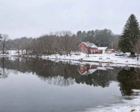 Red historic buildings and snow-dusted trees reflected in a calm pond at Blackstone River and Canal Heritage State Park during late February.