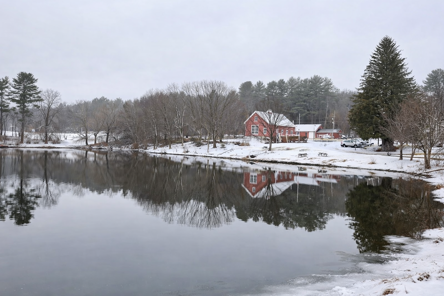 Red historic buildings and snow-dusted trees reflected in a calm pond at Blackstone River and Canal Heritage State Park during late February.