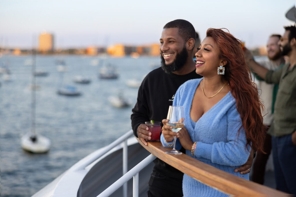 Couple enjoying cocktails on the deck of the Odyssey during a Boston Harbor dinner cruise with sailboats and the city skyline in the background.