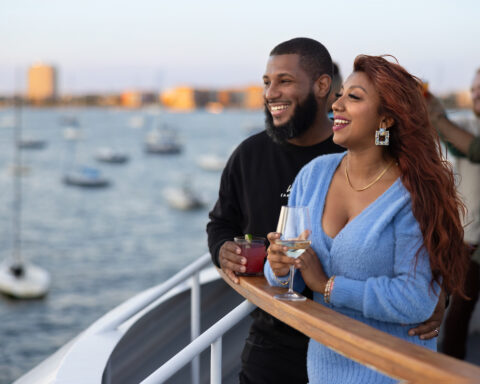 Couple enjoying cocktails on the deck of the Odyssey during a Boston Harbor dinner cruise with sailboats and the city skyline in the background.