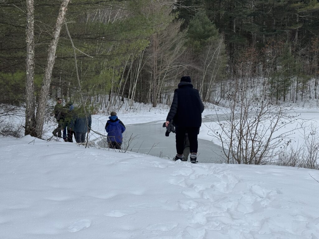 Adults and children walking through snow toward a partially frozen shoreline at Wachusett Reservoir in winter.
